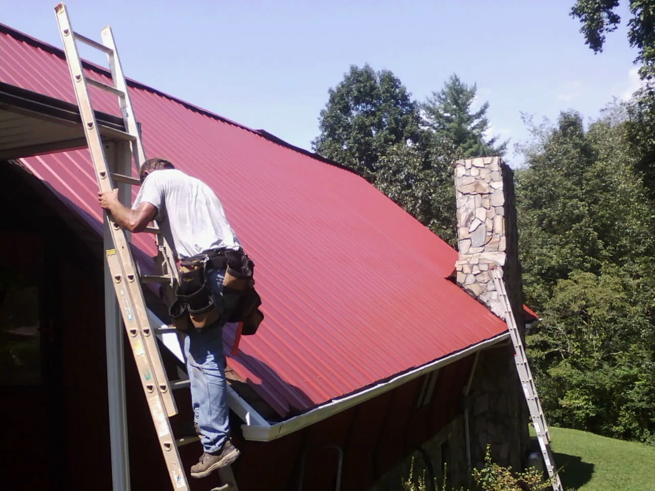 Roofer on a ladder beside a red metal roof and stone chimney — Soffit & Fascia Repair by Roofing Brothers in Essex Fells